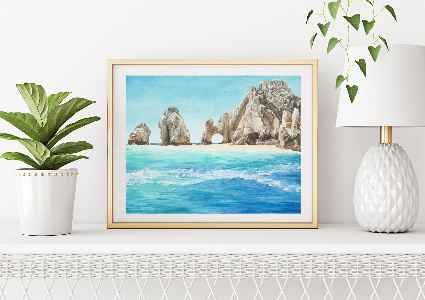 A framed El Arco fine art print displayed on a shelf with a plant to the left and a lamp above it, set against a white wall.