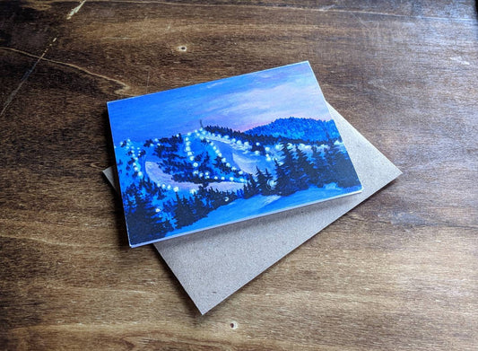 Square greeting card showing a nighttime mountain ski scene with glowing trail lights and evergreen trees, photographed on a wooden surface with a kraft envelope beneath.