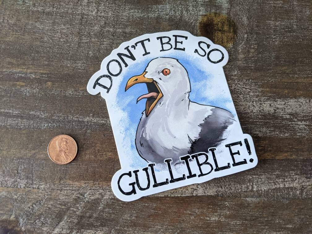 A sticker with an illustration of a seagull and the phrase 'DON'T BE SO GULLIBLE!' written on it, placed on a wooden surface, next to a penny for scale.