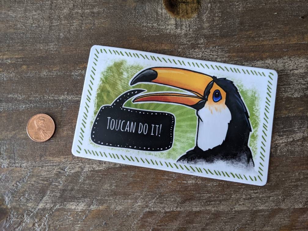 Toucan vinyl sticker with a speech bubble reading “Toucan do it,” shown next to a penny for size reference on a wooden surface.
