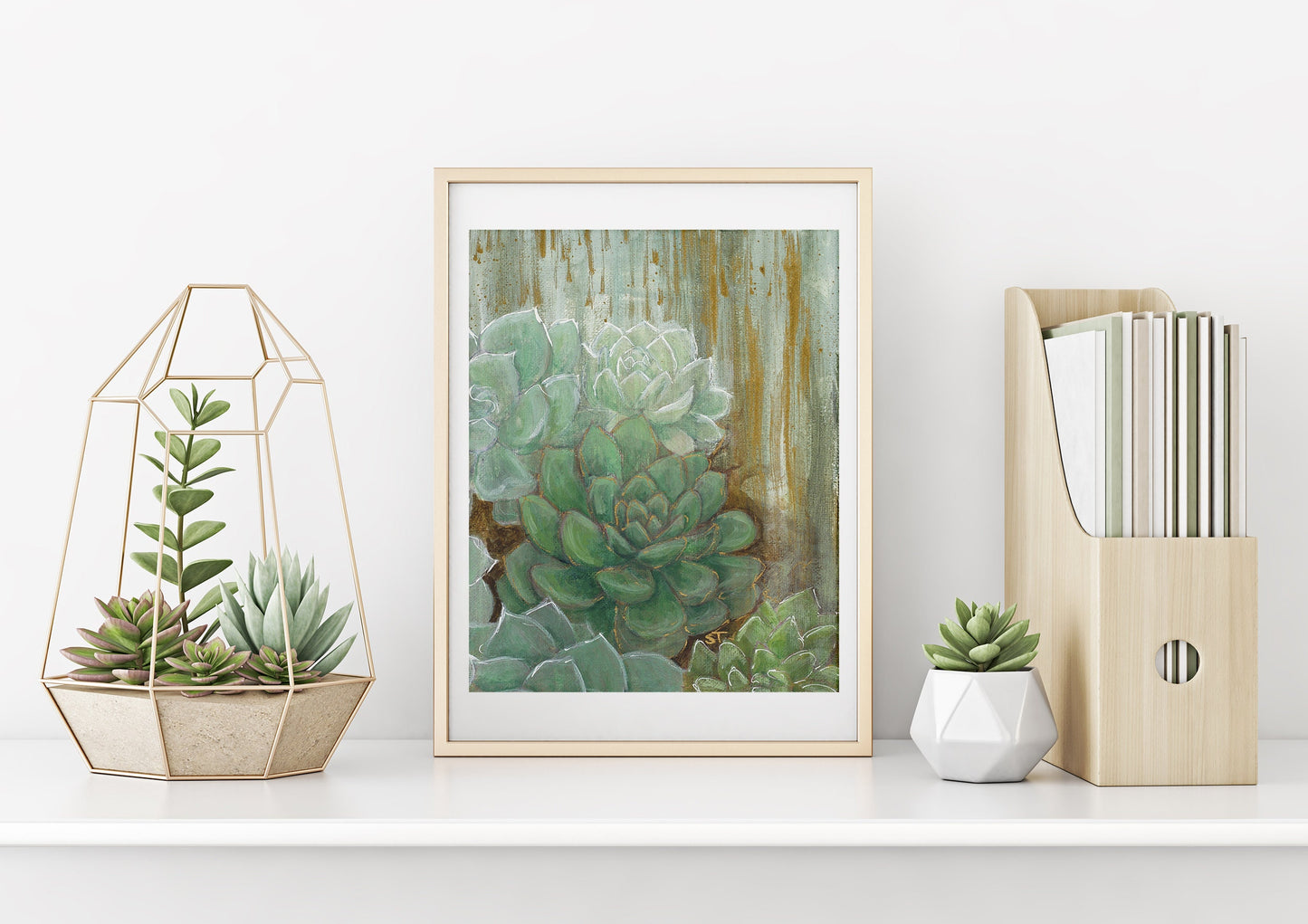 A framed art print featuring abstract green and gold colors with a depiction of succulent plants, placed on a shelf with a couple potted succulents and magazine holder.
