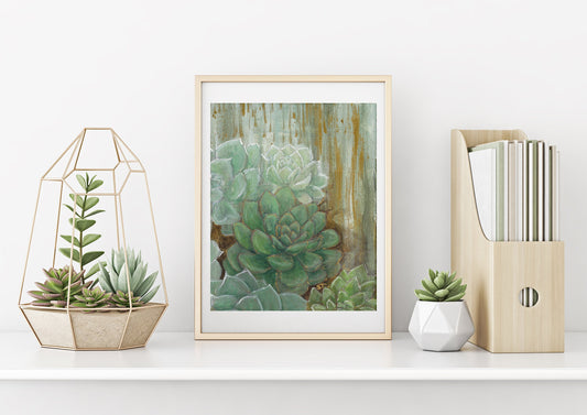 A framed art print featuring abstract green and gold colors with a depiction of succulent plants, placed on a shelf with a couple potted succulents and magazine holder.