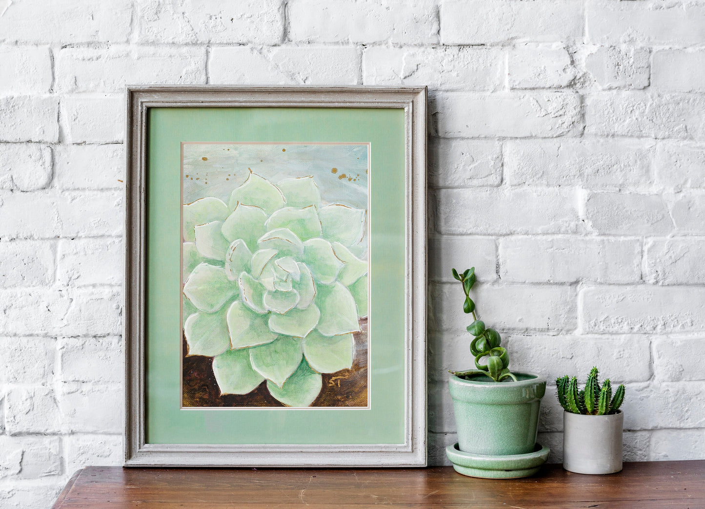 Framed pale green succulent art print by Sarah Terrell styled on a wooden surface against a white brick wall with small potted plants.