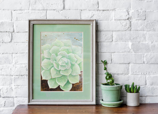 Framed pale green succulent art print by Sarah Terrell styled on a wooden surface against a white brick wall with small potted plants.