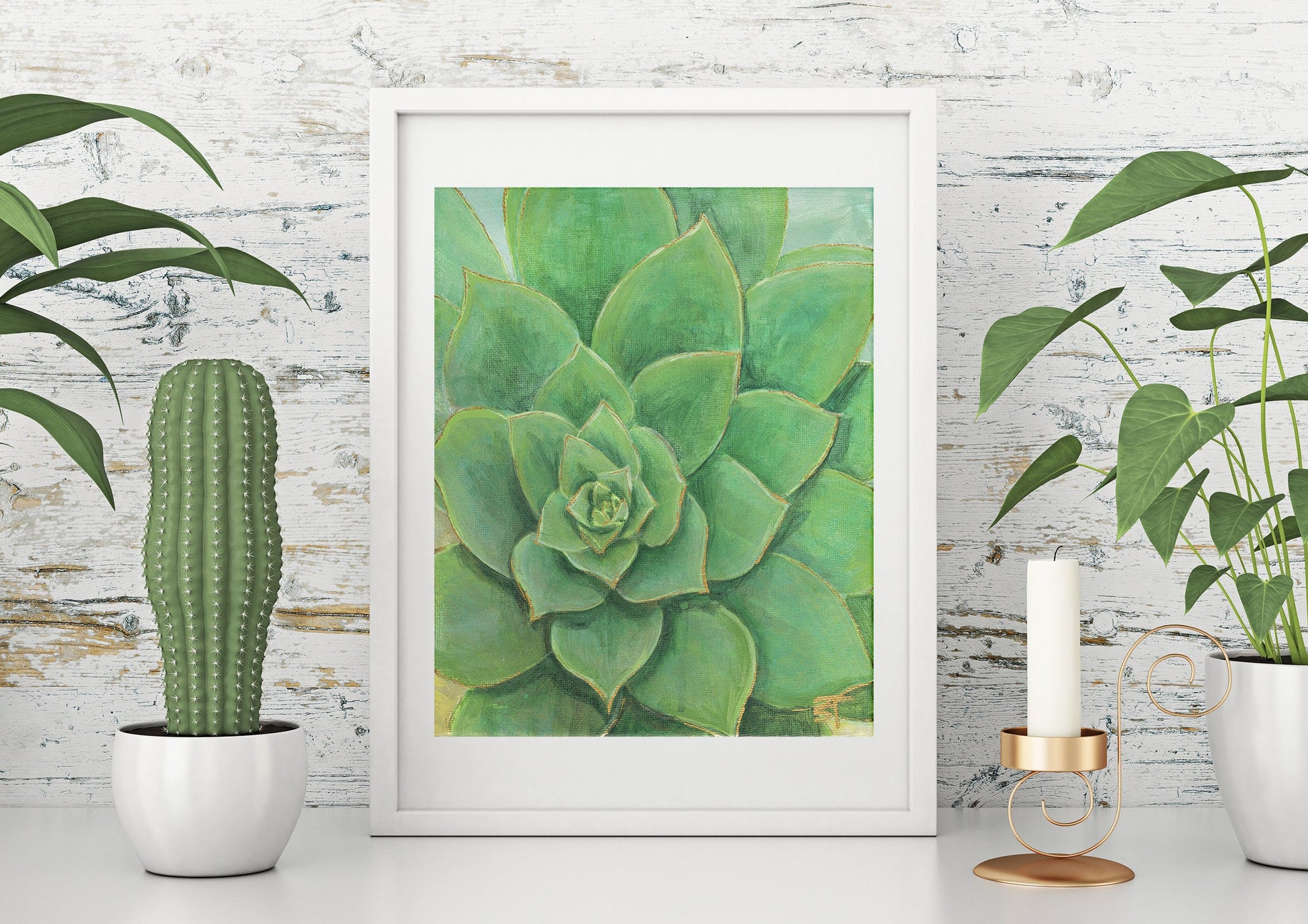A framed art print of a succulent with gold accents displayed on a white wall, flanked by potted plants and a candle.